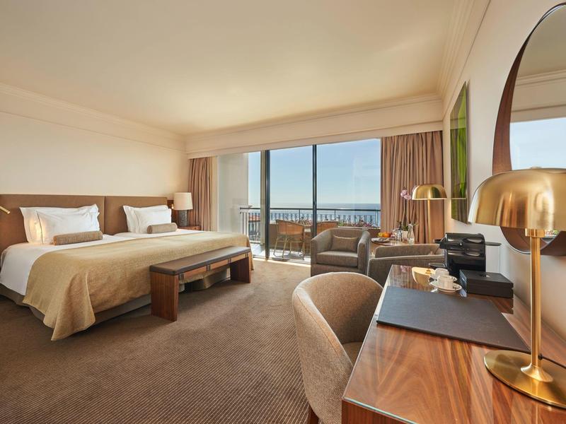 Camera d'hotel luminosa con letto matrimoniale, scrivania, sedia e vista mare attraverso grandi finestre.