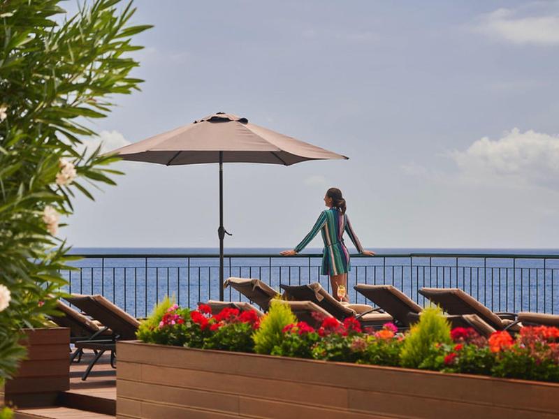 Terrazza con lettini e ombrellone con vista sul mare e piante fiorite.