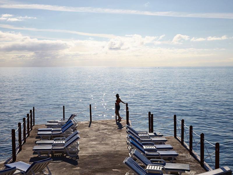 Persona in piedi alla fine di un molo con sedie a sdraio, che guarda il mare al tramonto.