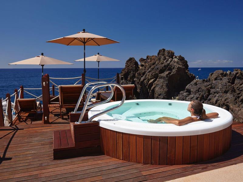 Donna si rilassa in vasca idromassaggio con vista sul mare e rocce, terrazza in legno con ombrelloni.