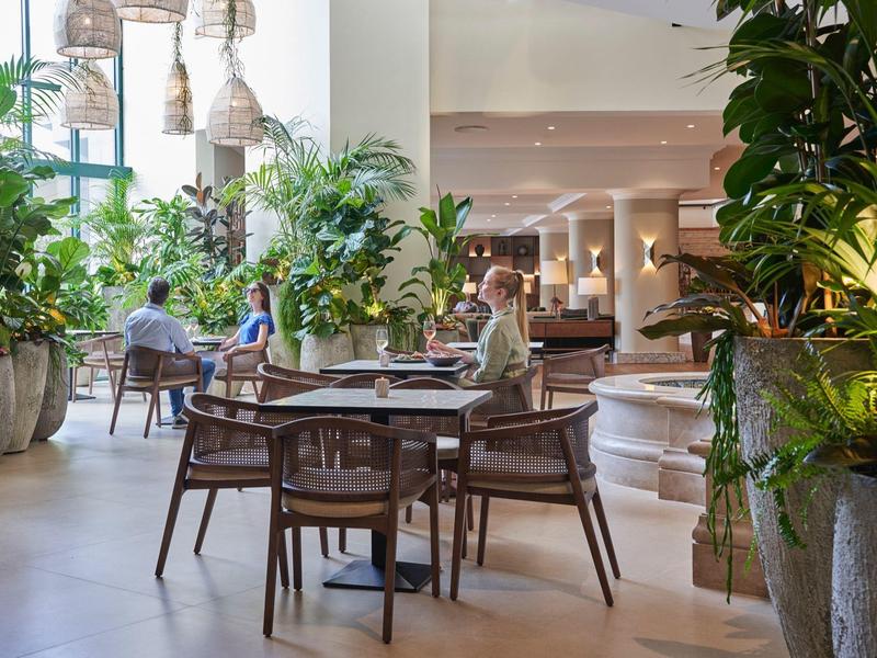 Area ristorante ben illuminata con tavoli in legno, sedie e molte piante verdi.