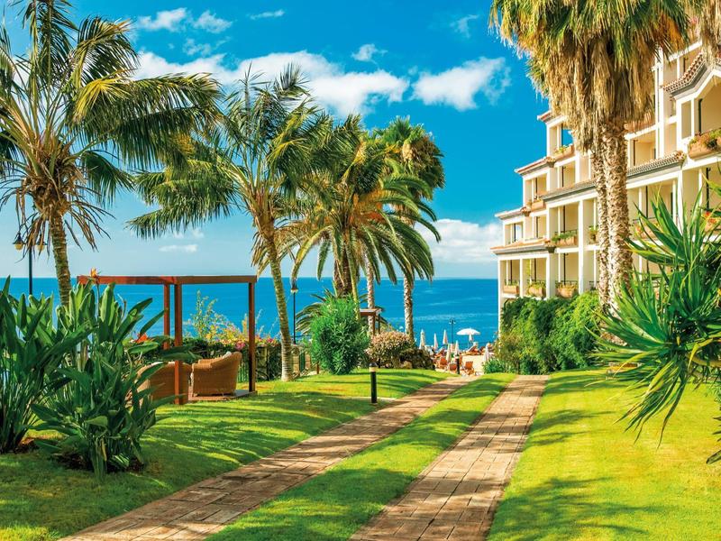 Sentiero con palme e piante che conduce a un hotel con vista mare sotto un cielo azzurro.