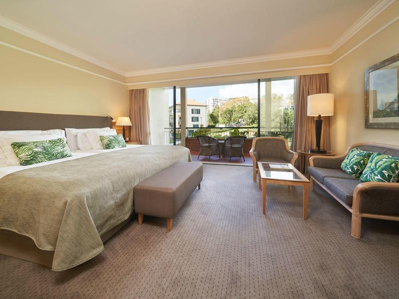 Camera d'hotel luminosa con letto king-size, divano, sedie e vista su piante verdi.