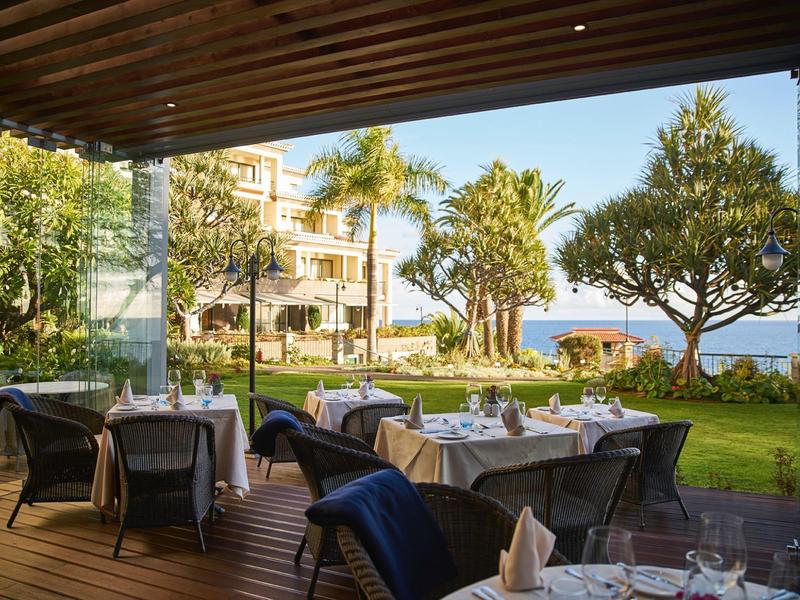 Terrazza con tavoli apparecchiati con vista su giardino e mare, circondata da palme e alberi.