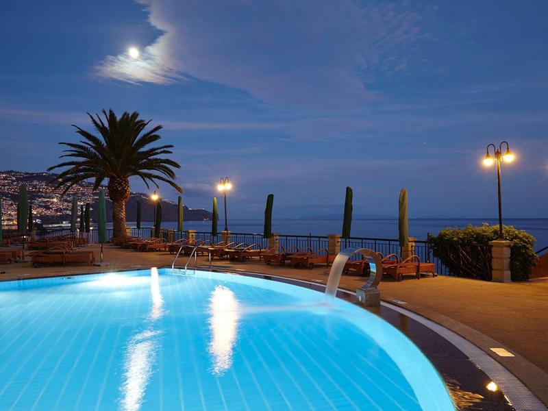 Piscina illuminata di notte con palme e mare sullo sfondo.