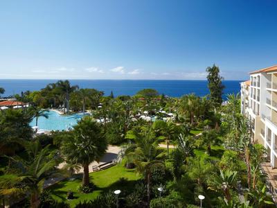 Pohled na hotelovou zahradu s bazénem, palmami a mořem v pozadí za jasného nebe.