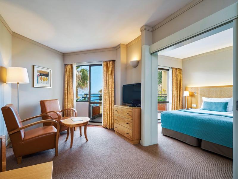 Světý hotelový pokoj s posezením, TV a přilehlou ložnicí s velkou postelí.