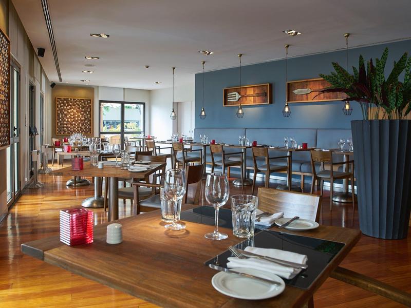 Moderní interiér restaurace s dřevěnou podlahou, prostřenými stoly a velkými rostlinami.