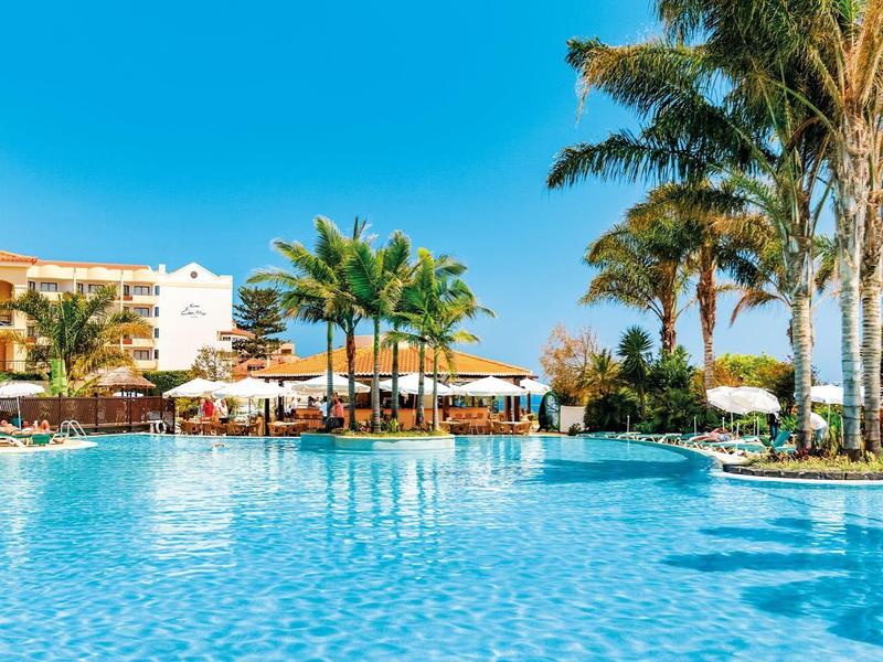 Velký bazén s čistou vodou, palmami a hotelovými budovami pod jasně modrou oblohou.