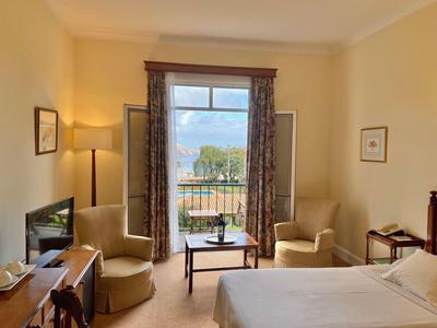 Helles Schlafzimmer mit Doppelbett, zwei Sesseln, kleinem Tisch, TV und Blick auf Balkon und Garten.