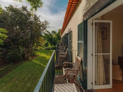Hausbalkon mit Holzstühlen, grünem Garten, Bäumen und offener Tür bei wolkigem Himmel.