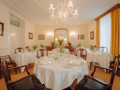 Elegantes Esszimmer mit rund gedeckten Tischen, Holzstühlen, Kronleuchter und großen Fensterläden.