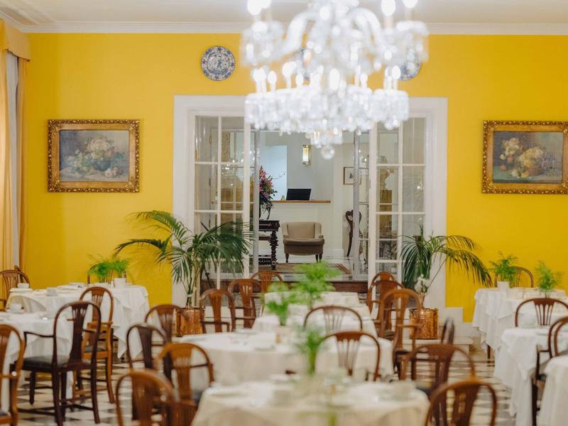 Hellgelber Speisesaal mit runden Tischen, weißen Tischdecken, Holzstühlen und Pflanzen im Raum.