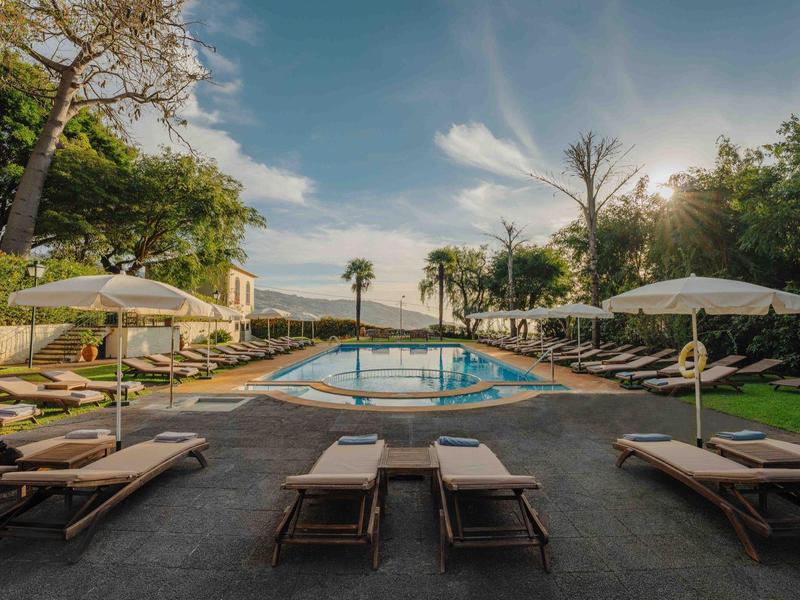 Outdoor-Pool mit Liegestühlen, Sonnenschirmen und grünen Bäumen unter blauem Himmel.