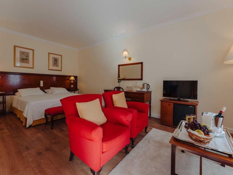 Hotelzimmer mit Doppelbett, roten Sesseln, Holzfußboden und Fernseher auf einem Sideboard.