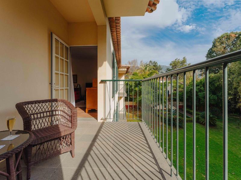 Balkon mit Stuhl, Tisch und Gläsern, Blick auf grünen Garten, sonniger Himmel mit Wolken.