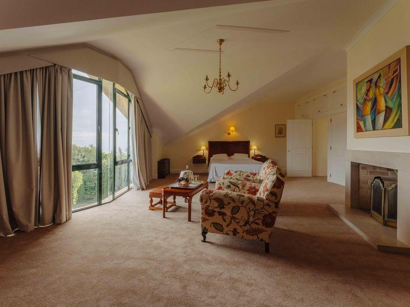 Großes Schlafzimmer mit schräger Decke, Kamin, Sofa, Sessel und Blick auf Garten durch Glasschiebetür.