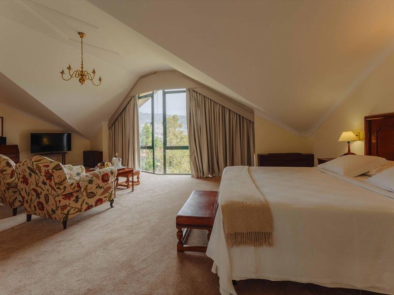 Ein gemütliches Schlafzimmer mit Doppelbett, zwei Sesseln, Fernseher und Blick aus großem Fenster.