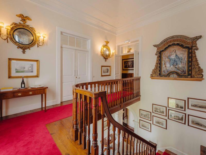 Heller Flur mit rotem Teppich, Holztreppe, antiken Spiegeln und gerahmten Bildern an den Wänden.