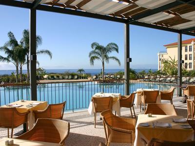 Freiluftrestaurant mit Holzstühlen, Tischen, Pool, Palmen und Meerblick unter blauem Himmel.