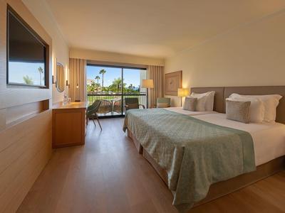 Habitación de hotel luminosa con cama doble, balcón y vista a palmeras a plena luz del día.