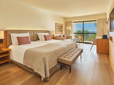 Helles Hotelzimmer mit zwei Betten, Holzmöbeln und Meerblick durch großes Fenster.