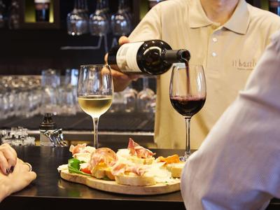 Ein Kellner füllt Rotwein in ein Glas, davor Vorspeisen und ein Glas Weißwein auf dunklem Tresen.