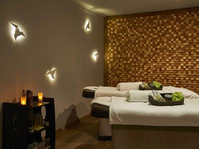 Ein ruhiger, gedämpft beleuchteter Wellnessraum mit zwei Behandlungsliegen und dekorativer Holzpaneelwand.