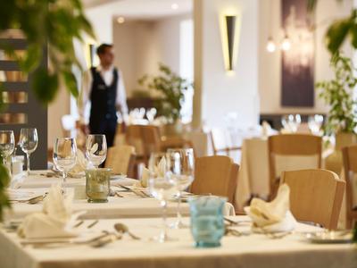 Eleganter gedeckter Tisch in hellem Restaurant mit Kellner im Hintergrund und dekorativen Pflanzen.