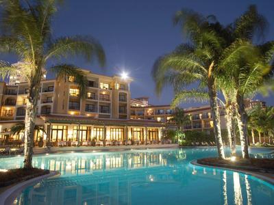 Hotel mit beleuchtetem Pool und Palmen vor mehrstöckigem Gebäude in der Abenddämmerung.