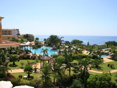 Blick auf einen Hotelgarten mit Palmen, Pool und Meer unter klarem blauem Himmel.