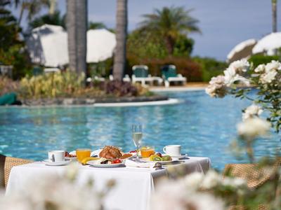 Tisch mit Frühstück am Pool, umgeben von Palmen und blauer Wasserfläche bei Sonnenschein.