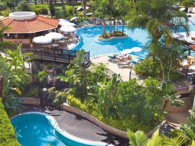 Blick auf tropischen Poolbereich mit Palmen, Sonnenschirmen und blauem Meer im Hintergrund.