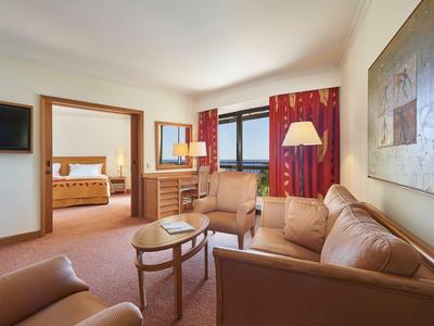 Helles Wohnzimmer mit braunem Sofa, Sesseln, Holztisch, rotem Vorhang und großem Spiegel.