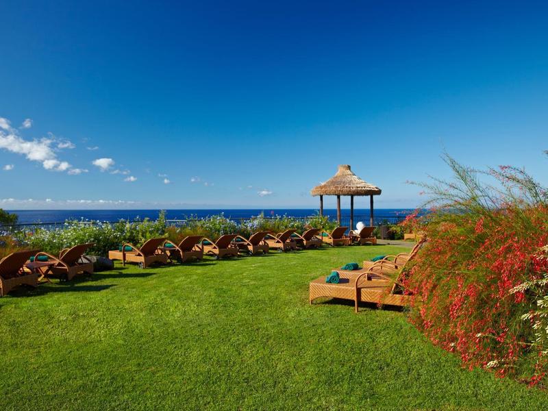 Césped verde con tumbonas y un quiosco mirando al mar azul bajo un cielo despejado.
