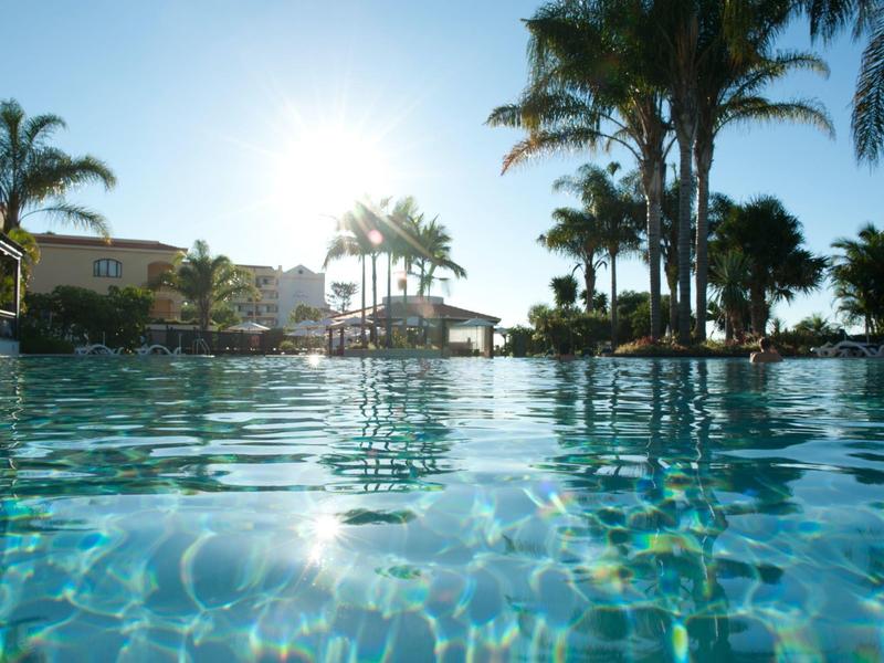 Klares Schwimmbad bei Sonnenaufgang mit Palmen und Hotelgebäuden im Hintergrund.