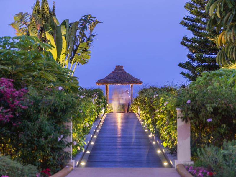 Pasillo iluminado conduce a un pabellón, rodeado de abundante vegetación y plantas al atardecer.