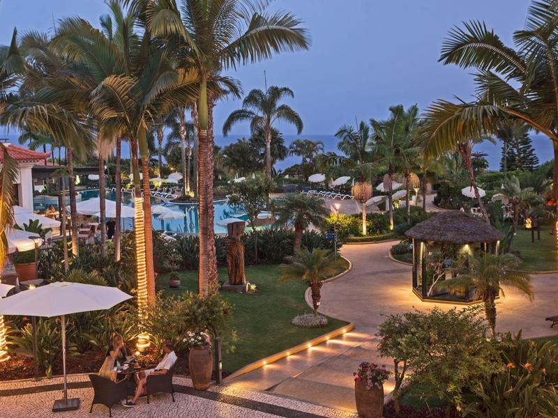 Área de resort con caminos iluminados, palmeras y mesas bajo sombrillas al atardecer