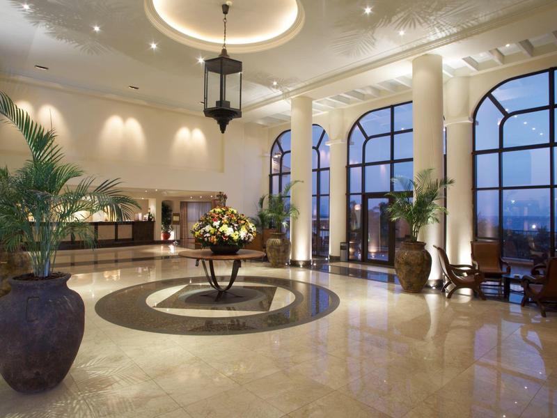 Amplio vestíbulo de hotel con grandes ventanas, plantas decorativas y suelo de mármol elegante al anochecer.