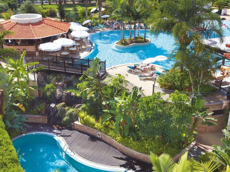 Vista de una piscina de resort tropical con palmeras, sombrillas y el mar al fondo.