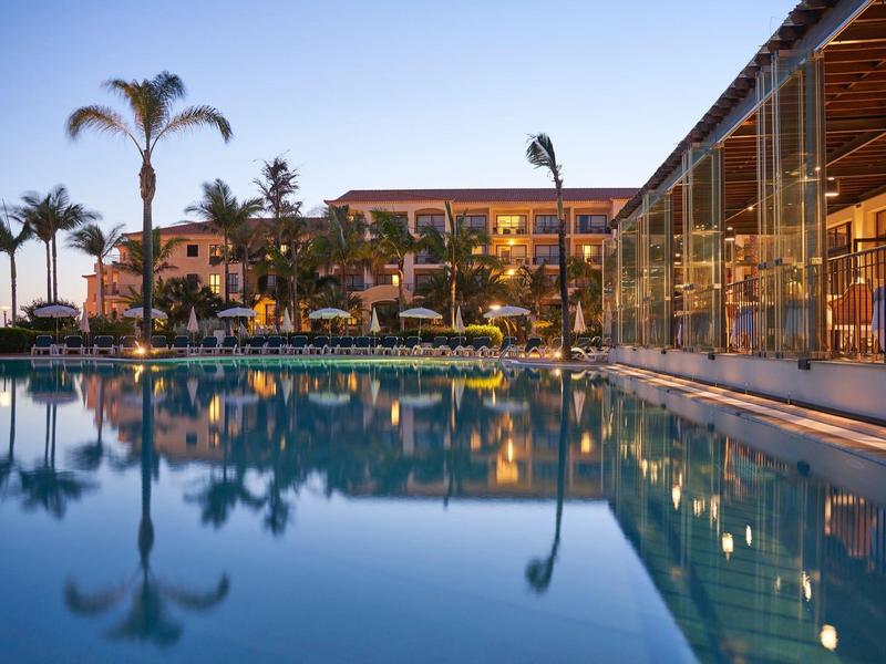 Piscina moderna de hotel al anochecer con palmeras y luces cálidas al fondo.
