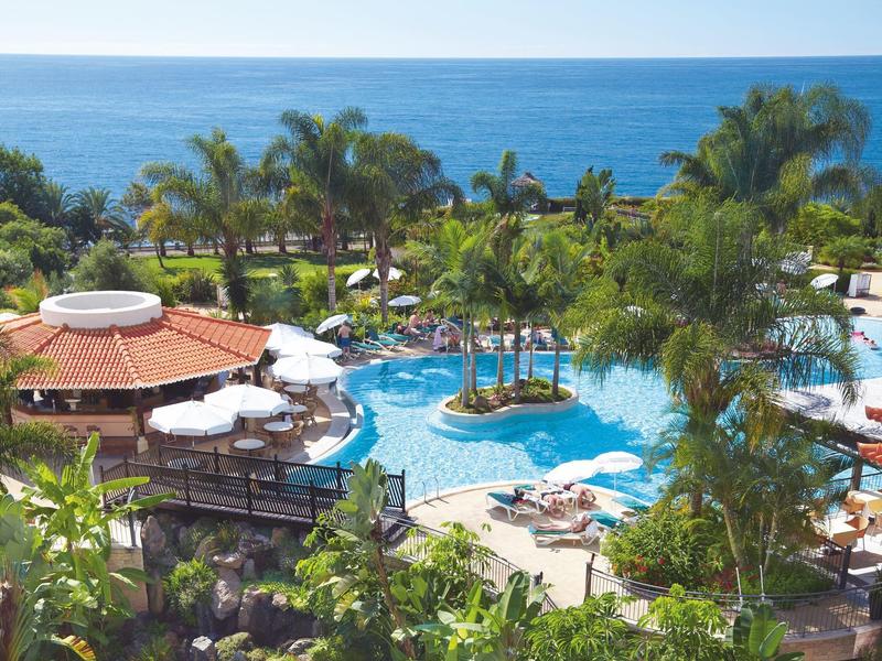 Área de piscina de resort con palmeras, sombrillas y vista al mar en día soleado.