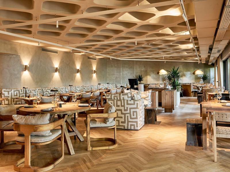Área de comedor de hotel con muebles de madera y iluminación cálida junto a ventanales grandes