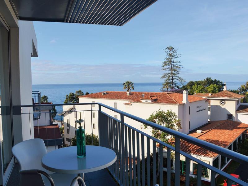 Balkon z stołem, krzesłami i widokiem na morze ponad białymi domami i niebieskim niebem.