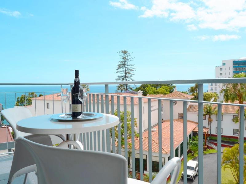 Balkon z białymi krzesłami, stołem, butelką wina i widokiem na morze w tle.