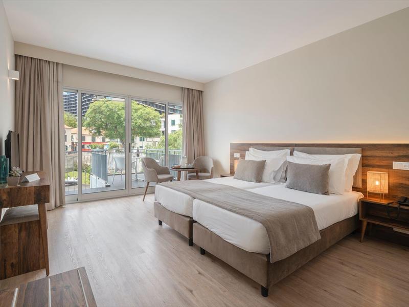 Jasny pokój hotelowy z podwójnym łóżkiem, stolikami nocnymi i widokiem na balkon.