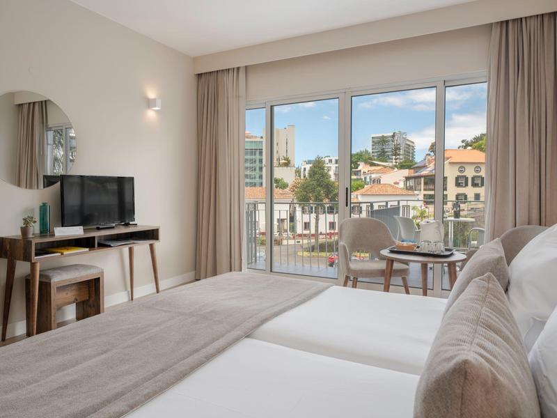 Jasny pokój hotelowy z wygodnym łóżkiem, telewizorem, biurkiem i balkonem z widokiem na miasto.