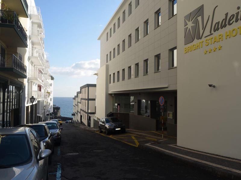 Widok na wąską ulicę z zaparkowanymi samochodami i budynkami, morze w tle.