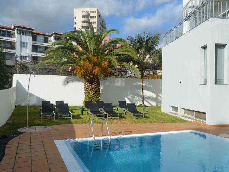 Mały basen z palmami i leżakami przed nowoczesnymi budynkami w słoneczny dzień.