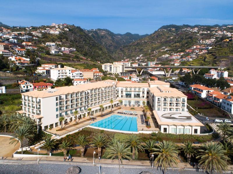 Hotel z dużym basenem otoczony budynkami i górami w tle pod niebieskim niebem.